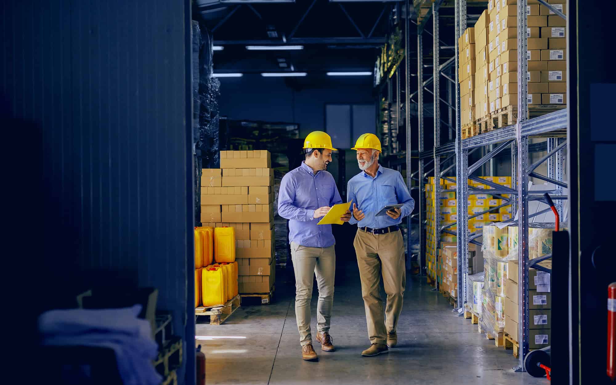 Two warehouse managers in hard hats reviewing paperwork between racks of imported goods